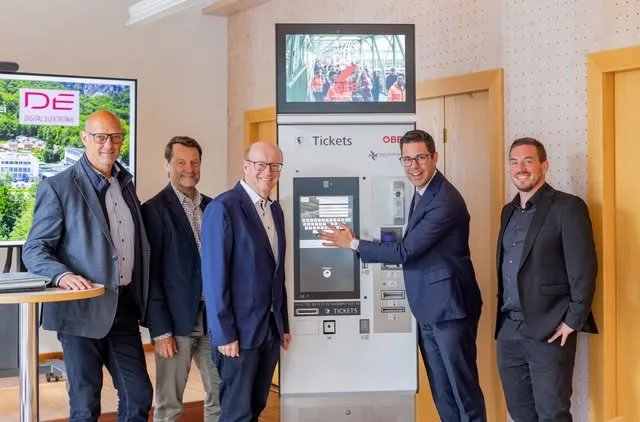 Mehrere Männer in Anzügen stehen neben einem ÖBB-Ticketautomaten, an der Wand ein Bildschirm mit dem DE-Logo