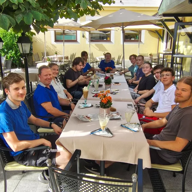 Lange Tischrunde junger Leute und Mitarbeiter in blauen Polos in einem Gastgarten