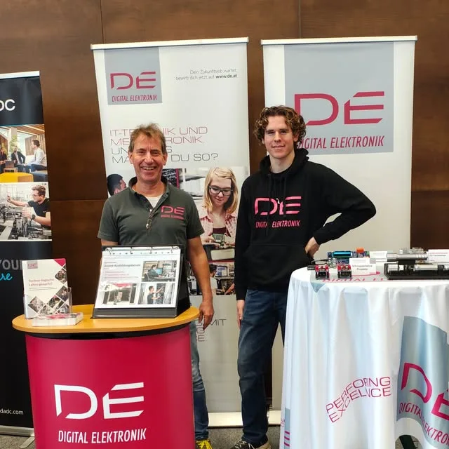Zwei Männer an einem Messestand mit Rollups und Pult im Magenta der Marke