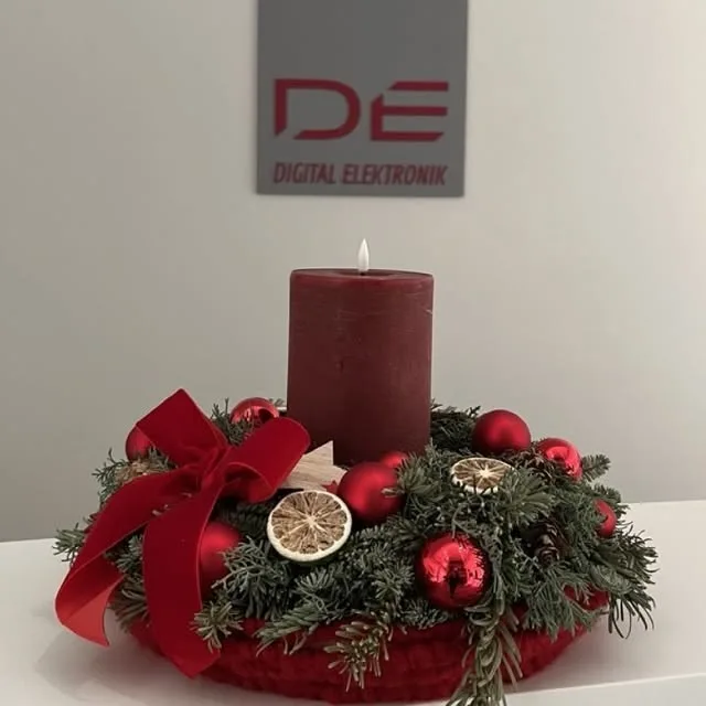Adventkranz mit roter Kerze auf einem Tisch, dahinter das DE-Logo an der Wand
